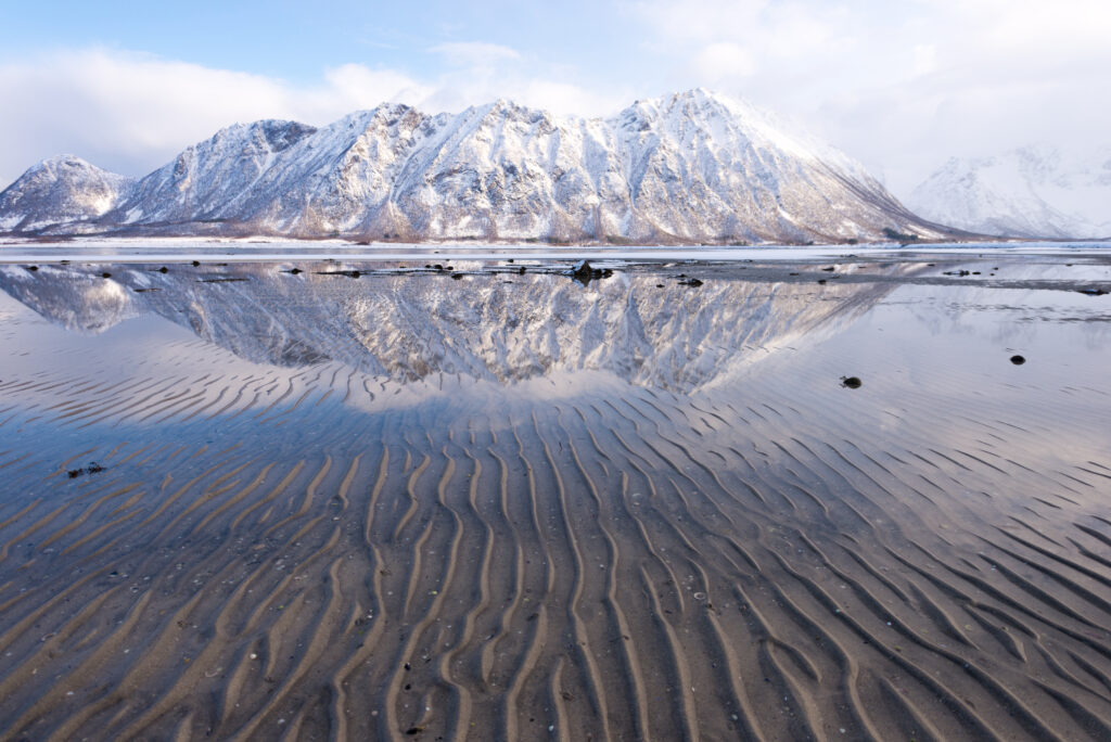 Snow-covered mountains reflected in calm water with rippled sand foreground winter wallpaper
