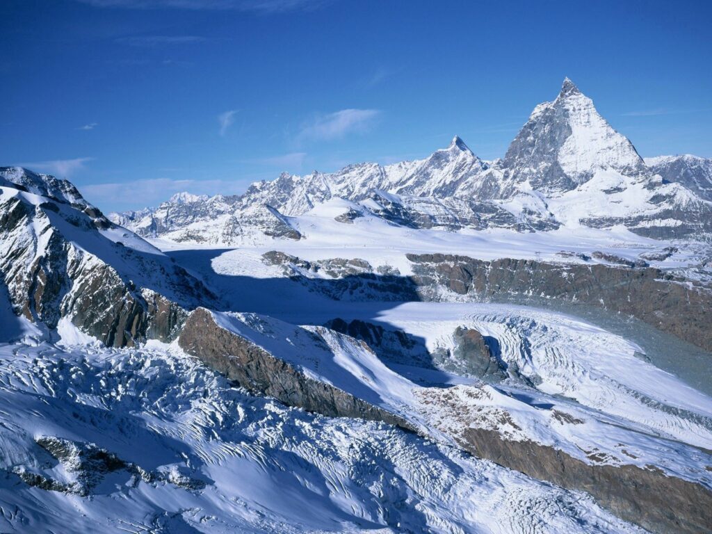Snow-covered mountains and rocky formations under clear blue sky in a serene winter landscape Wallpaper