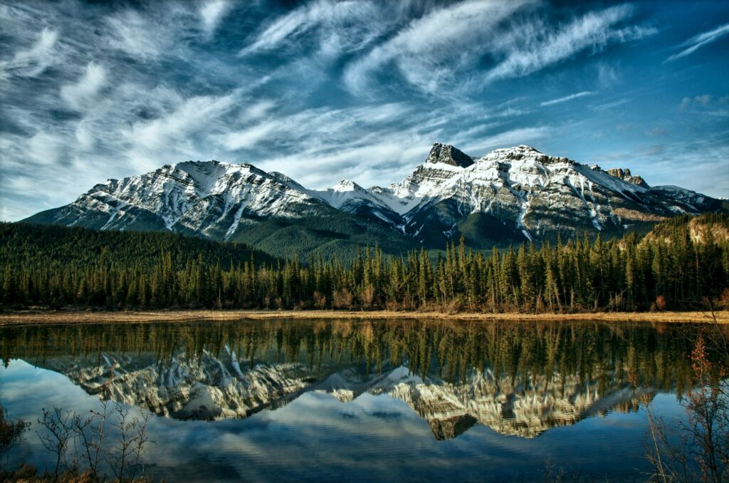 Snow-capped Canadian mountains and pine forests reflected in calm lake with vibrant sky background Wallpaper