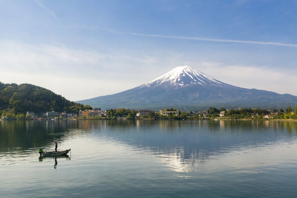 Snow-capped mountain reflecting on calm lake with boat and lush greenery under blue sky Wallpaper