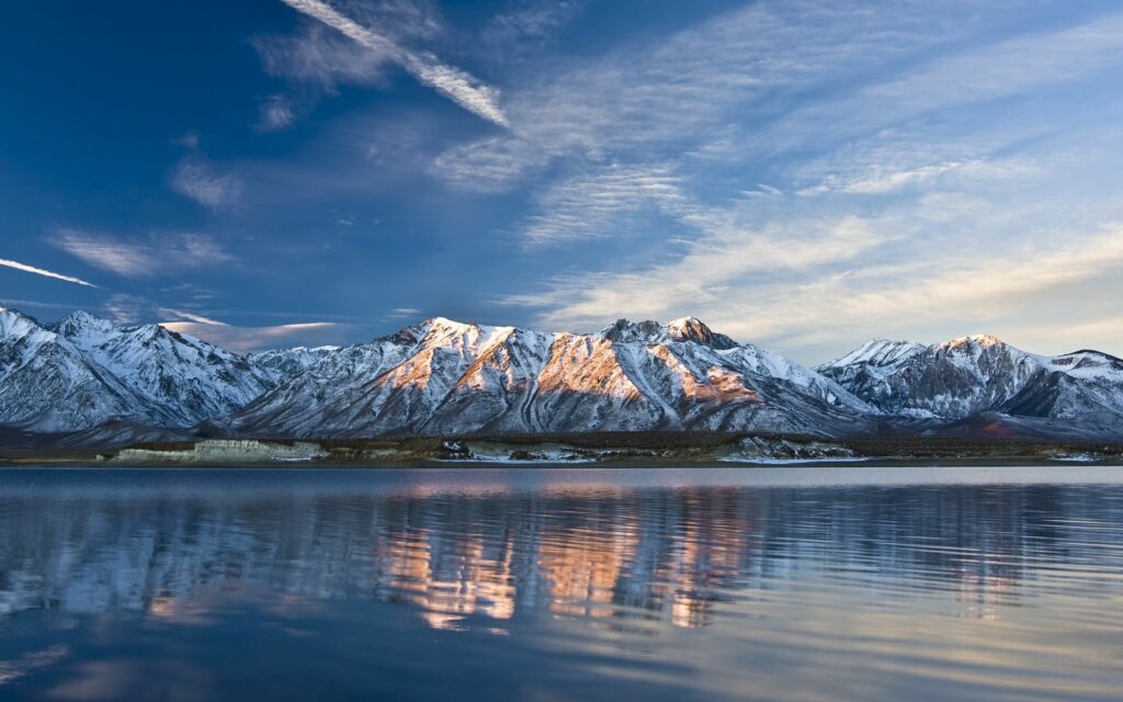 Serene snow-capped mountains reflected in a calm lake with soft sunlight and wispy clouds Wallpaper