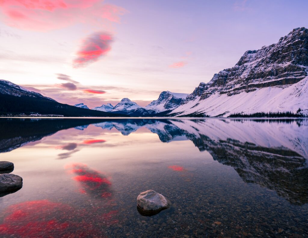 Snow-capped mountains reflected in clear water at sunset with pink clouds and rocky foreground Wallpaper