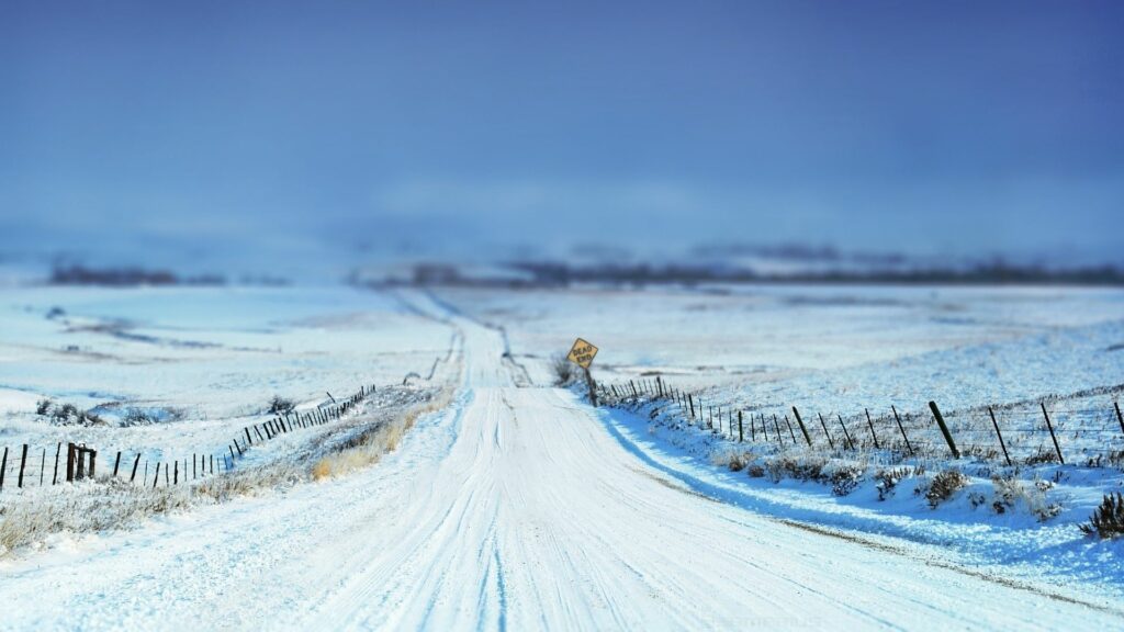 Snowy winter road with yellow sign bordered by stakes and fences across white fields wallpaper