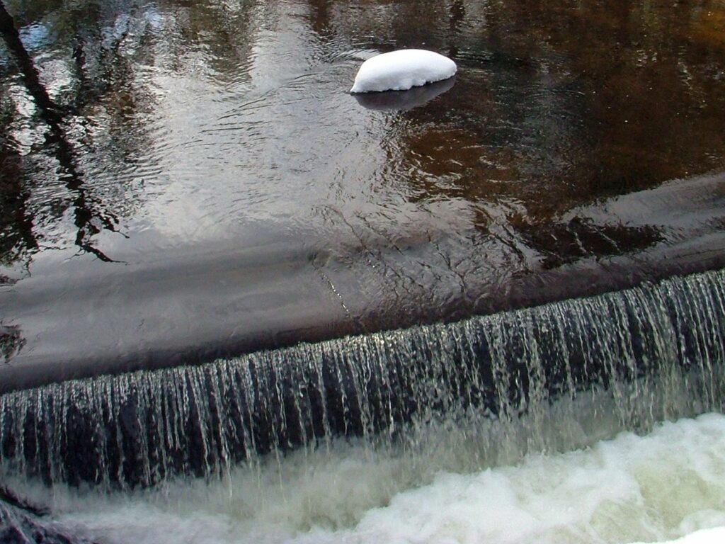 Serene river cascading over dam with clear water and snow patch reflecting early spring light Wallpaper