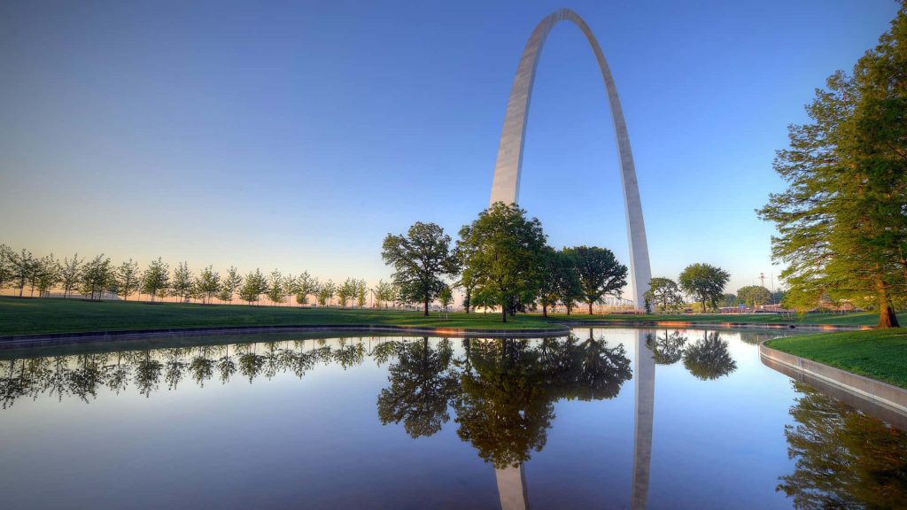 St Louis Gateway Arch reflected in calm pond with greenery and soft blue sky background wallpaper