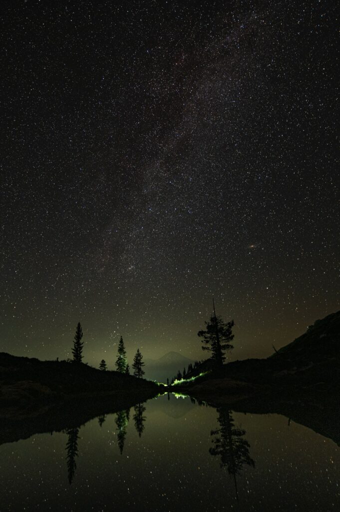 Serene night sky with Milky Way and stars reflected in calm lake with silhouetted trees and mountain Wallpaper