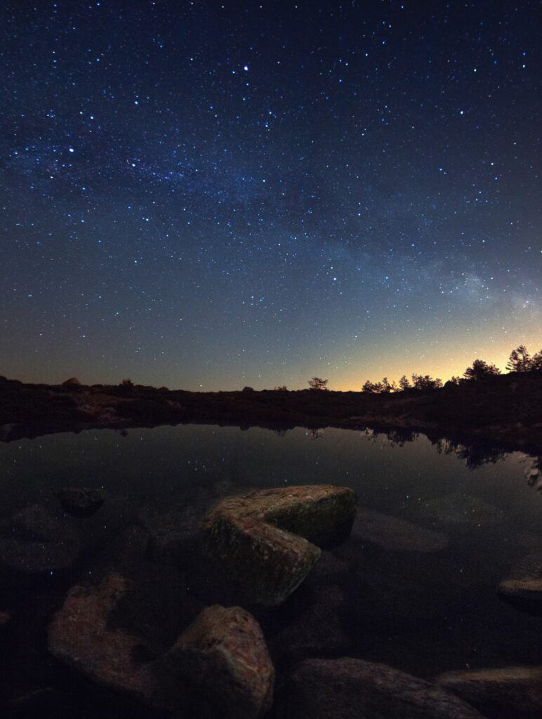 Serene starry night sky reflects on calm lake with distant trees and foreground rocks tranquil scene Wallpaper