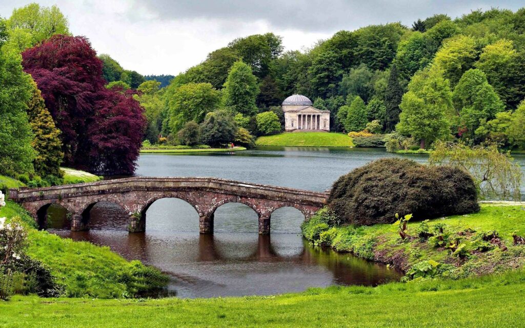 Serene landscape with classic stone bridge over calm river surrounded by lush trees and pavilion Wallpaper