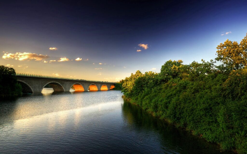 Stone bridge with arches over calm river surrounded by green trees at sunset wallpaper background
