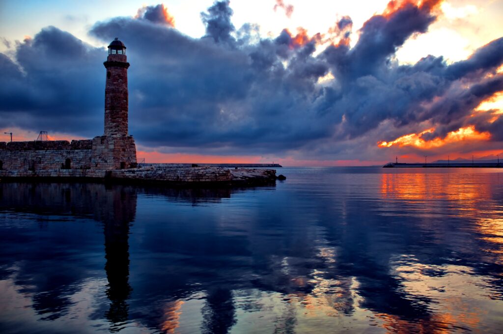 Stone lighthouse on historic pier reflected in calm waters at colorful sunset sky wallpaper background