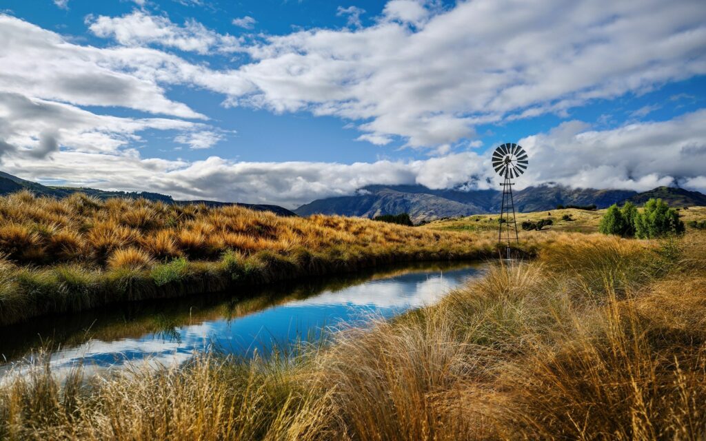 Serene summer landscape with reflective river golden grass blue sky rustic windmill wallpaper background