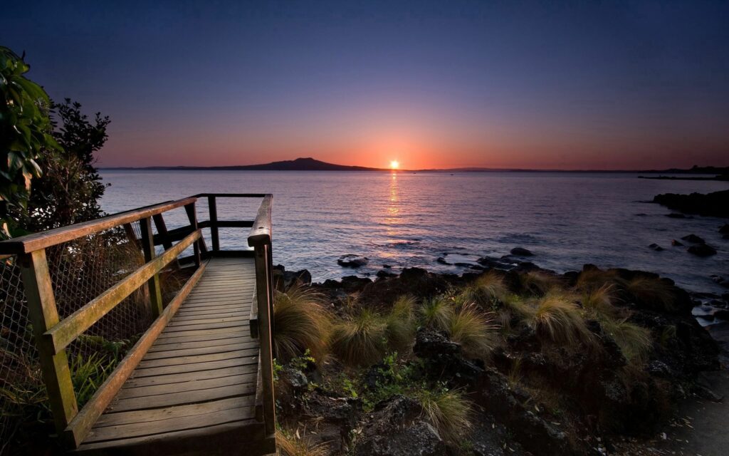 Sunset over calm lake with tall grasses wooden walkway and mountain silhouette summer sky Wallpaper