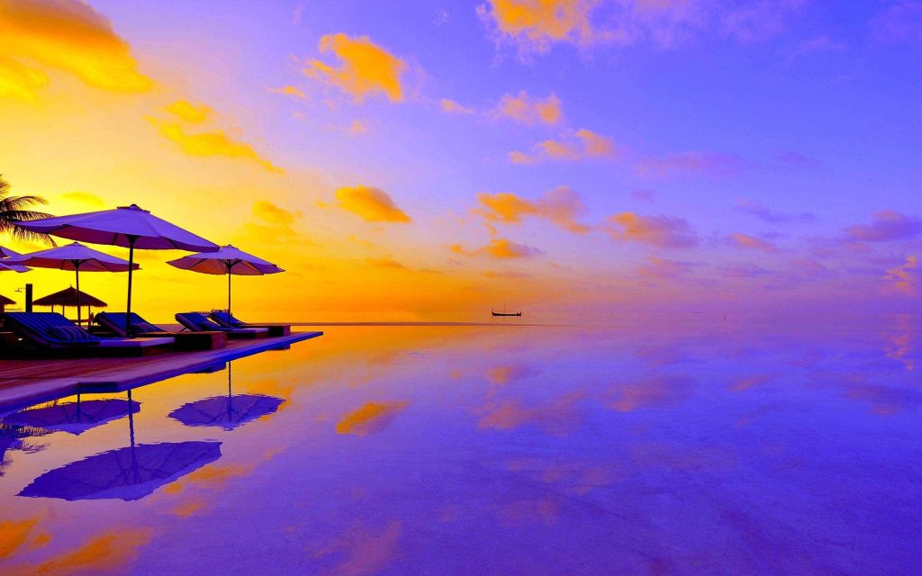Luxury resort infinity pool at sunset with beach loungers and purple orange sky background Wallpaper