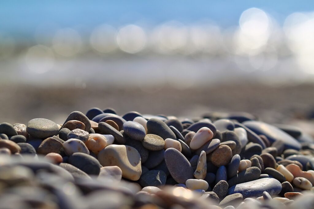 Smooth earth tone pebbles on sunlit beach with blurred blue ocean and sky serene wallpaper background