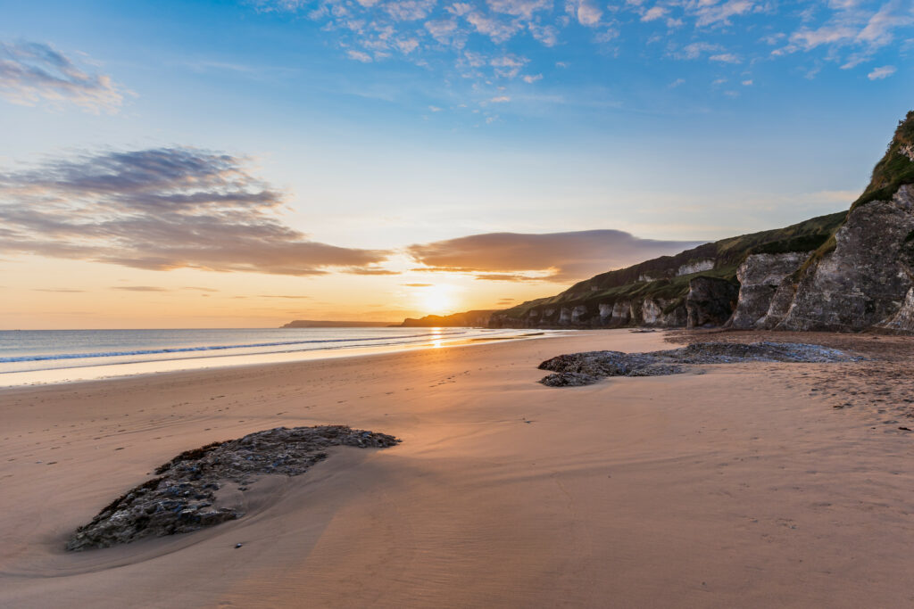 Sunrise beach scene with golden light sandy shore smooth stones rocky cliffs calm sea horizon Wallpaper