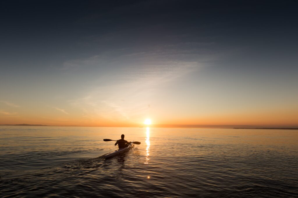 Man paddling boat on calm sea at golden sunrise with distant skyline peaceful morning scene Wallpaper