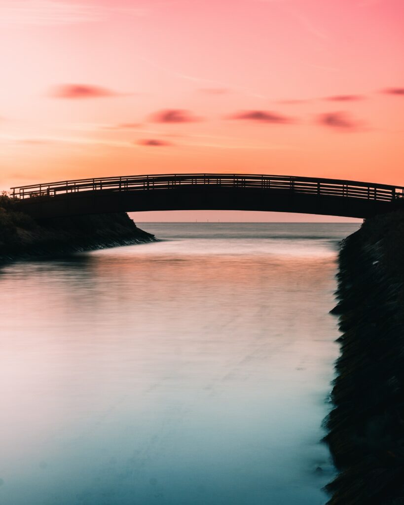 Serene bridge over calm bay at sunset with pink orange sky reflecting on tranquil water wallpaper