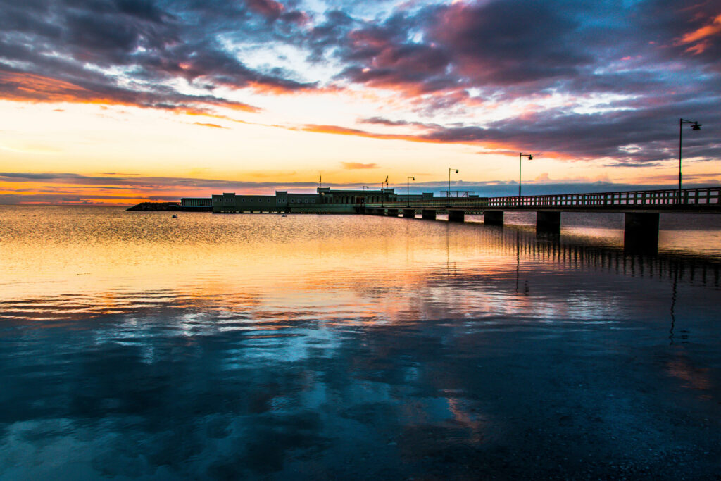 Serene sunset with long bridge over calm sea reflecting vibrant orange pink and blue hues Wallpaper