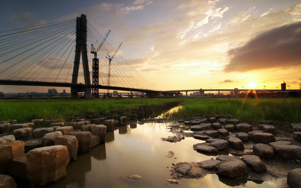 Sunset cityscape with modern cable-stayed bridge over calm river and green grass foreground Wallpaper