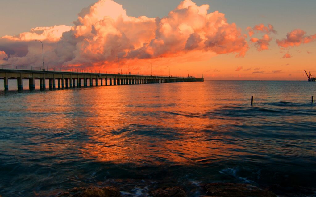 Tranquil dock at sunset over calm sea with vibrant orange and pink clouds reflecting water Wallpaper
