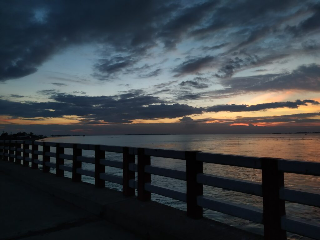 Serene seaside embankment at sunset with orange blue sky reflecting on calm water and railing Wallpaper