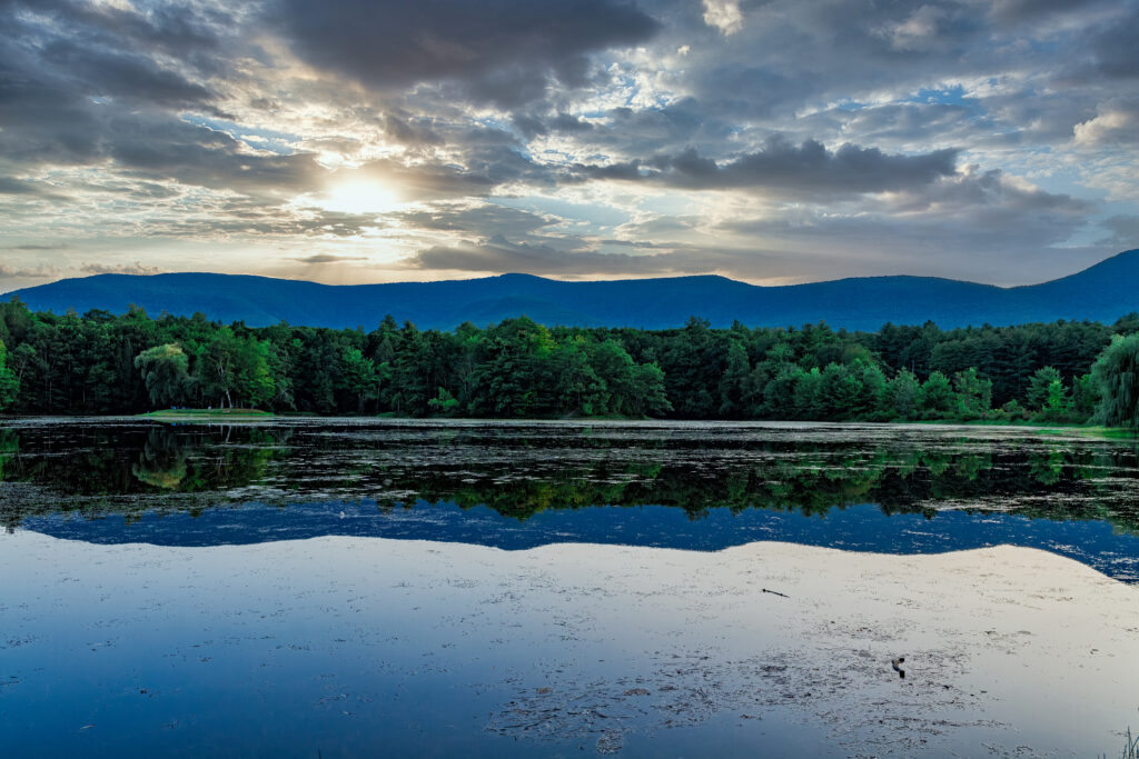 Sunset over calm pond reflecting dense forest lush trees distant mountains and dramatic sky Wallpaper