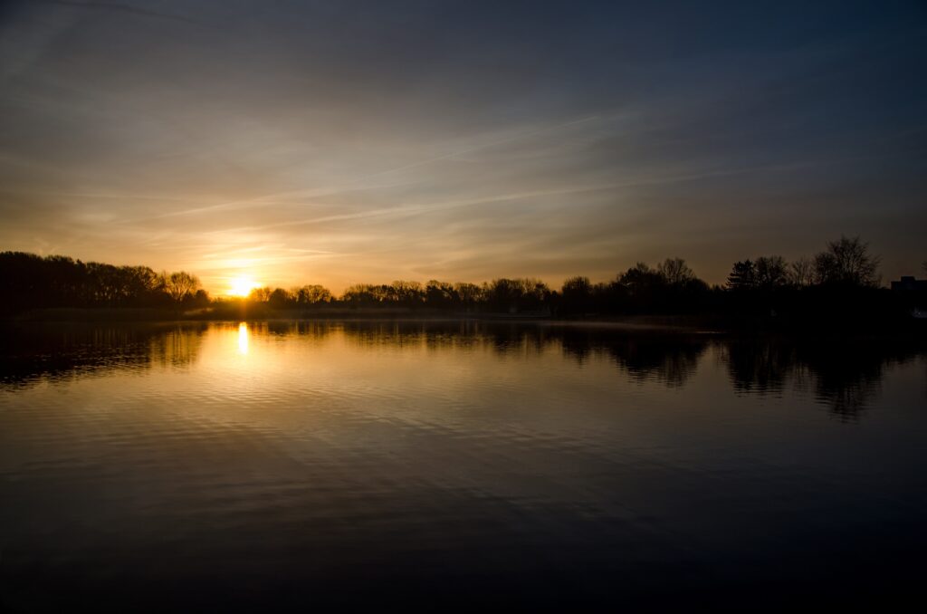 Sunset over calm lake with warm sky hues and silhouetted trees reflecting on tranquil water wallpaper