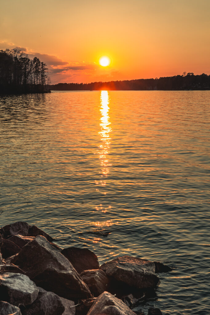 Serene lake at sunset with vibrant orange sky calm water reflecting dense forest and rocks foreground Wallpaper