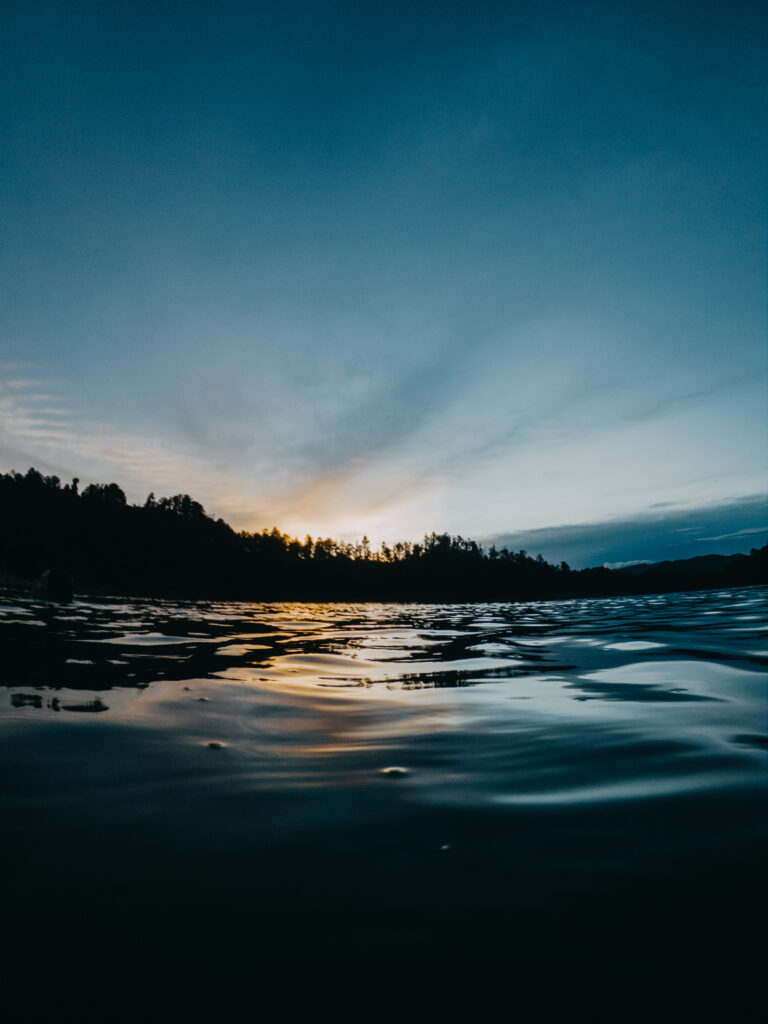 Serene lake sunset with rippling water dark tree-lined shore and dramatic orange blue sky background Wallpaper