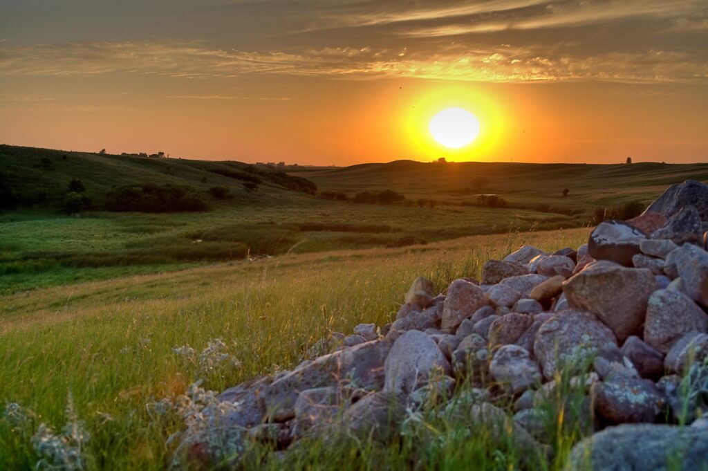 Serene sunset over rolling green fields with glowing sun and weathered stone pile foreground wallpaper