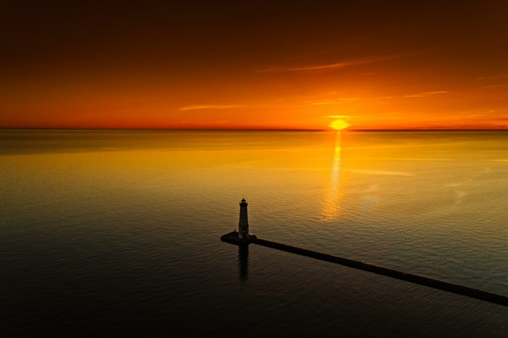 Aerial view of solitary lighthouse at sunset with orange sky reflecting on calm sea waters wallpaper