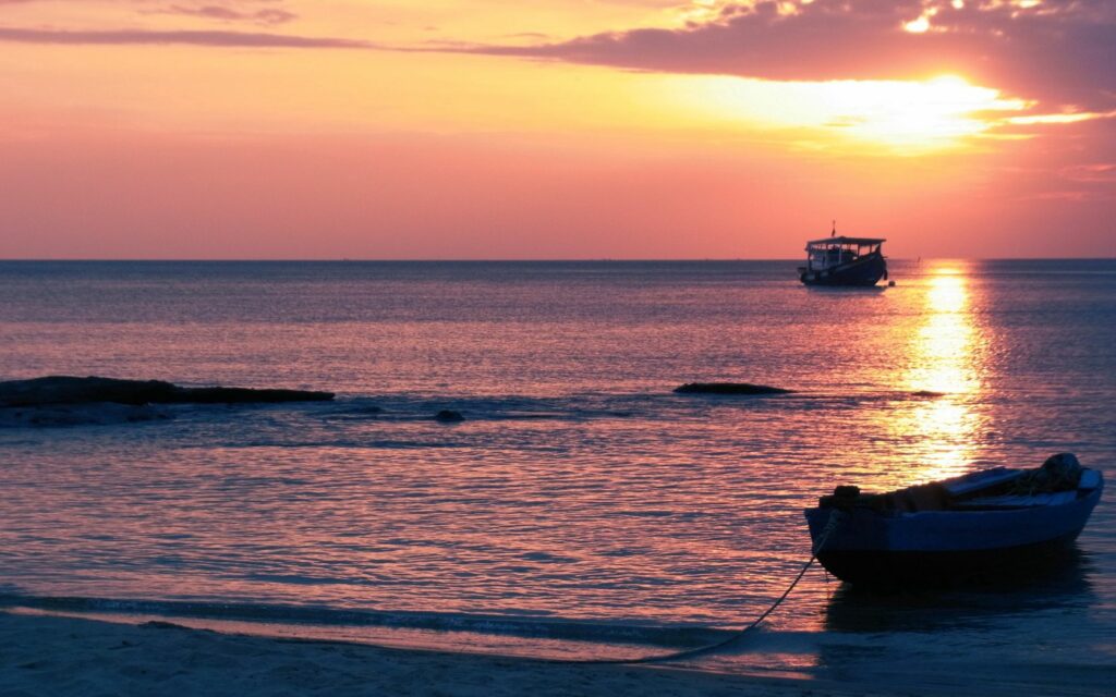Two boats float peacefully on calm ocean at sunset with warm orange pink sky and reflections Wallpaper