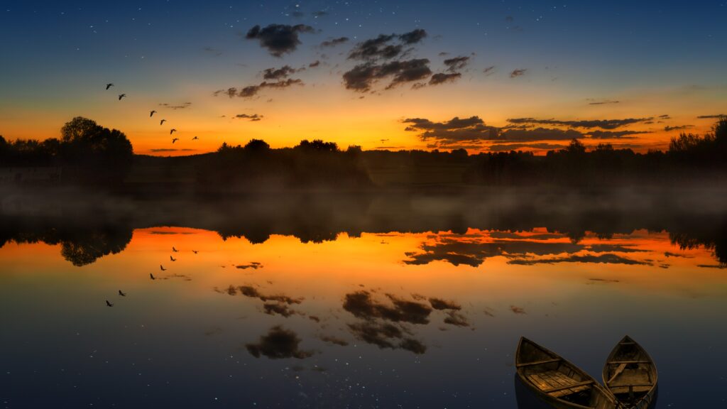 Serene sunset over calm lake with orange blue sky reflections and two boats in foreground Wallpaper