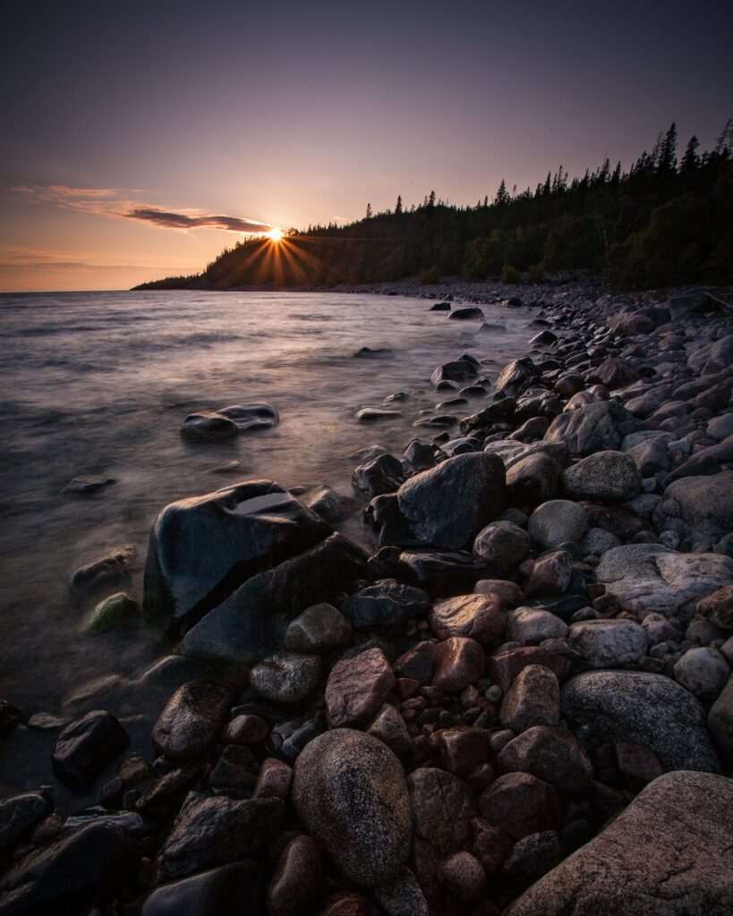 Sunset over calm sea with gentle waves smooth stones lush trees silhouette golden light wallpaper