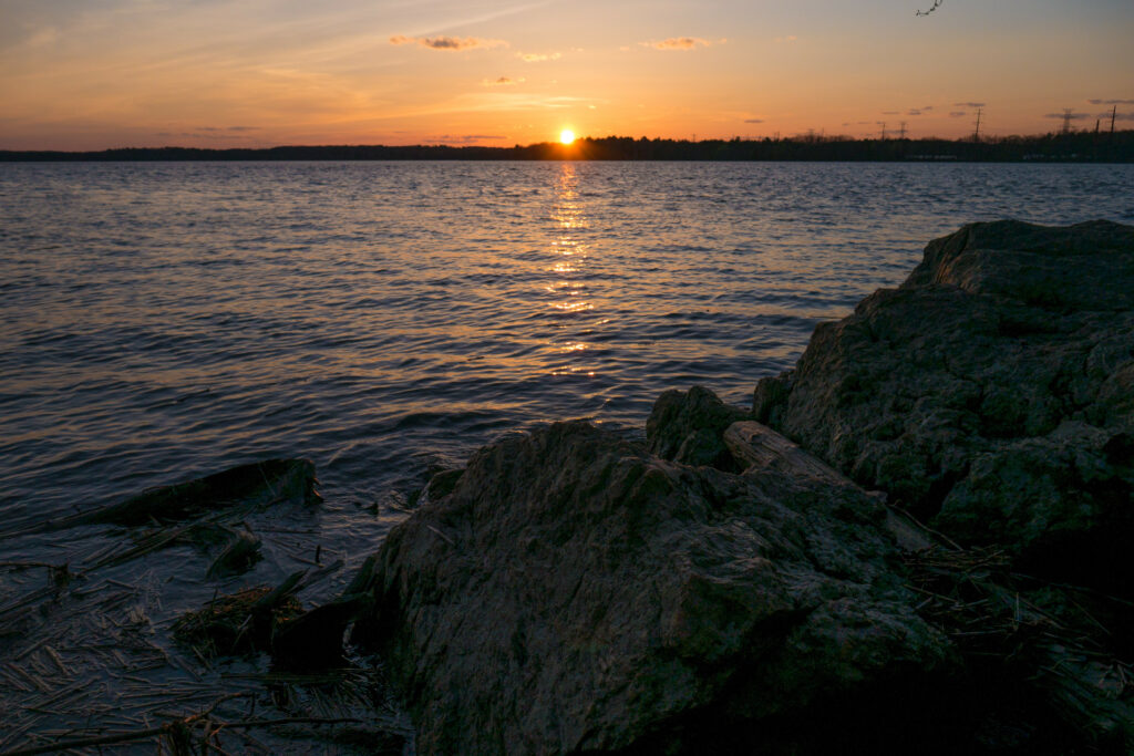 Glowing sunset over calm water with orange reflections and rugged rocks in foreground wallpaper