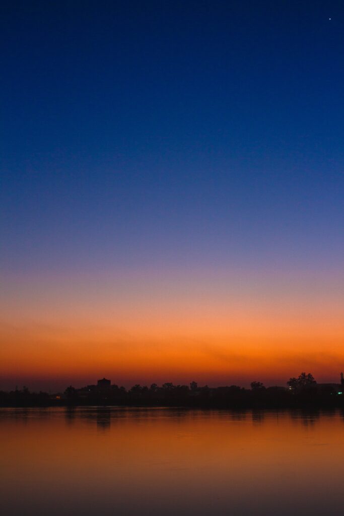 Serene sunset over calm water with vibrant gradient sky and silhouetted trees on shore Wallpaper