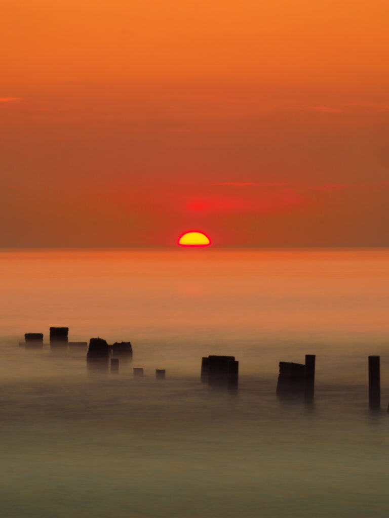 Serene coastal sunset with warm orange hues foggy sea and wooden posts creating dreamy wallpaper