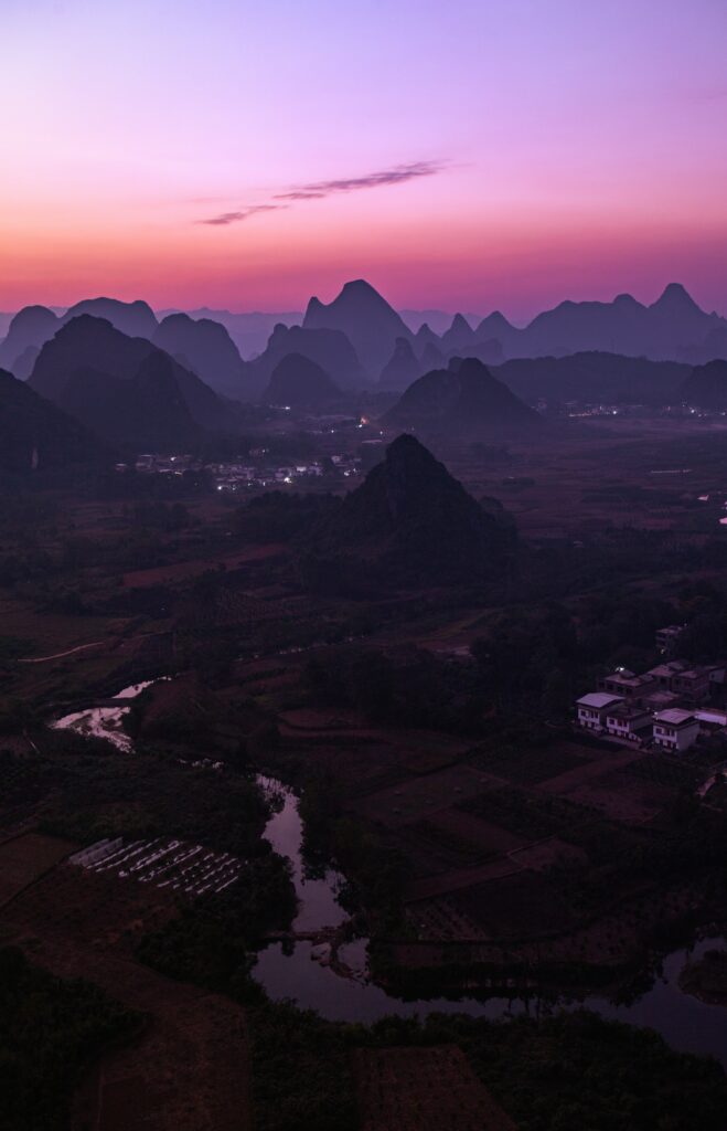 Serene sunset over mountains with quaint buildings fields and rivers in calming purple and orange hues Wallpaper
