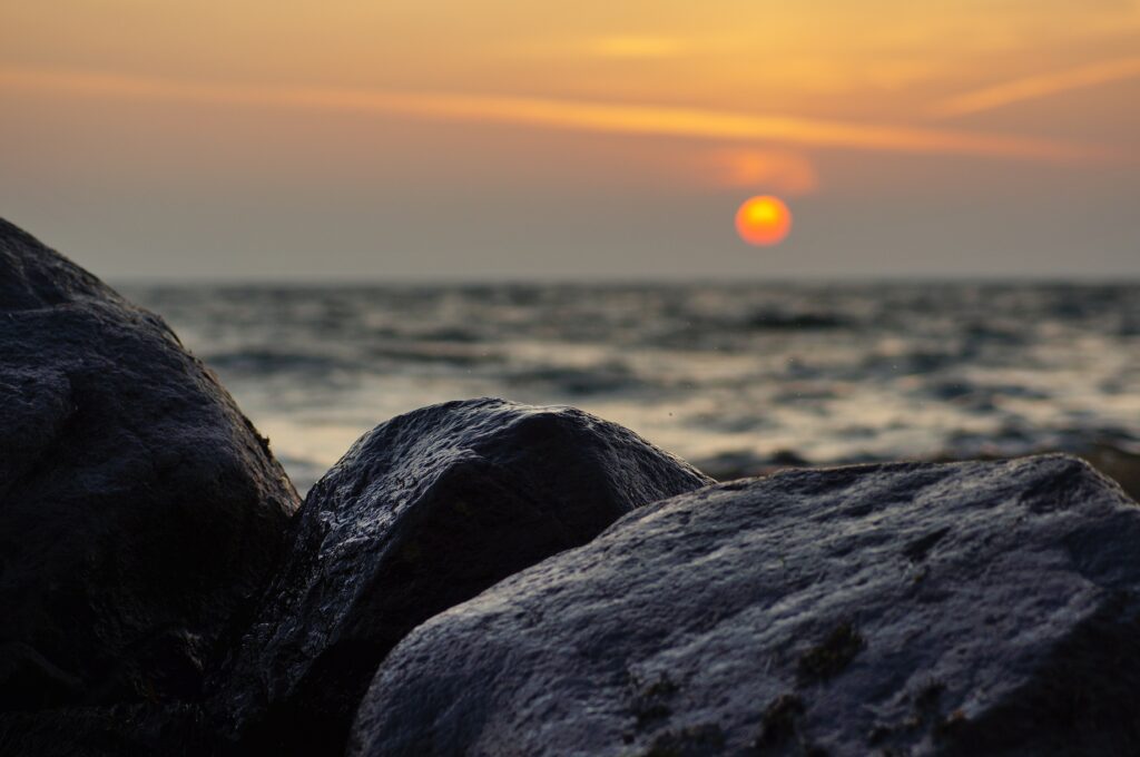 Serene sunset over calm sea with glowing orange sun and blurred coastal rocks foreground wallpaper