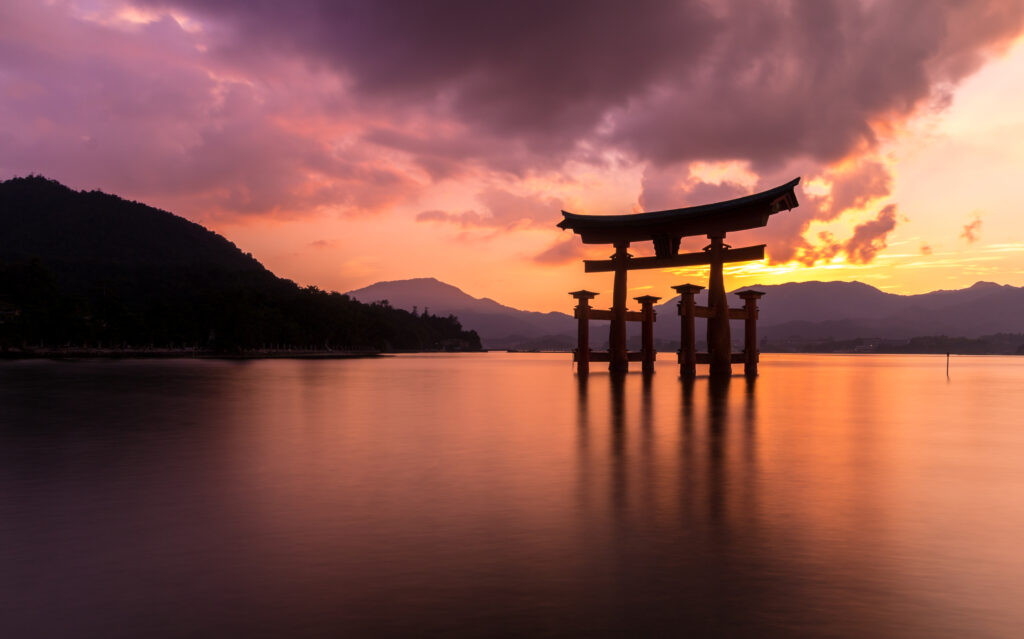 Sunset over tranquil sea with silhouetted torii gate dramatic clouds and distant mountains wallpaper