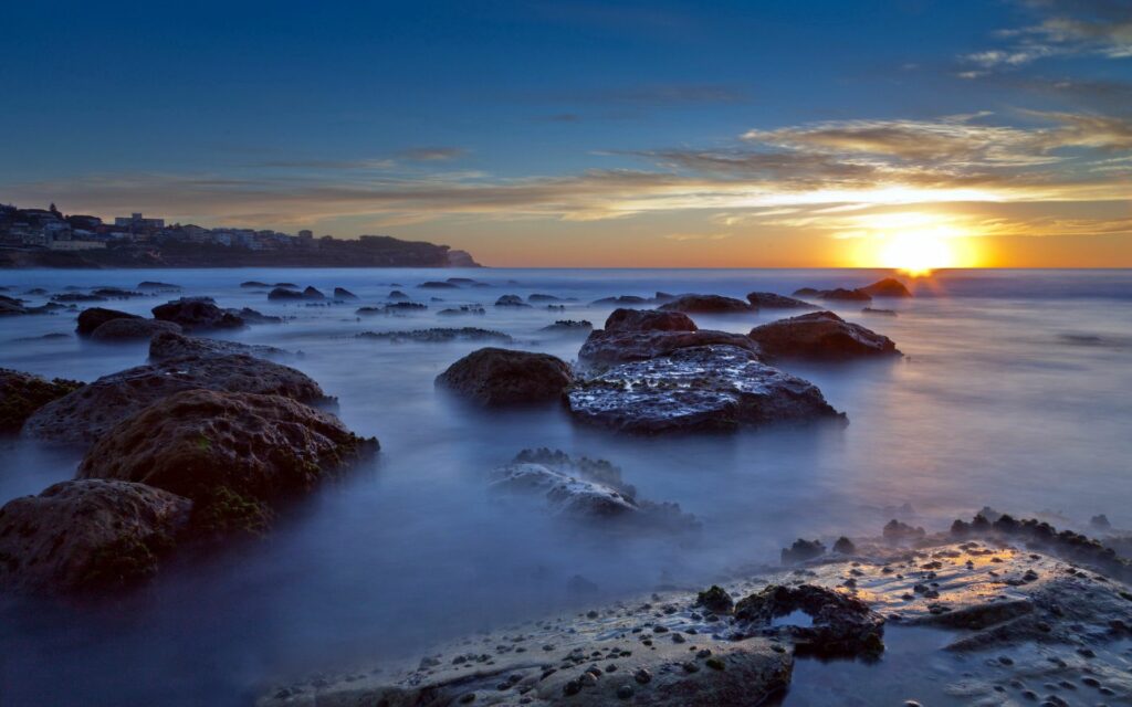 Serene sunset over calm sea with golden light reflecting on waves and dark coastal rocks Wallpaper