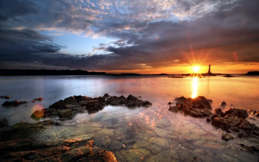 Evening sea scene with clear water around rocks and a distant beacon under a sunset sky Wallpaper