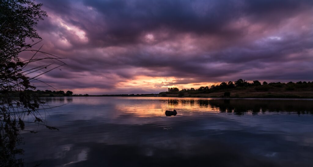 Serene river at sunset with tree silhouettes calm water and dramatic cloud-filled twilight sky wallpaper