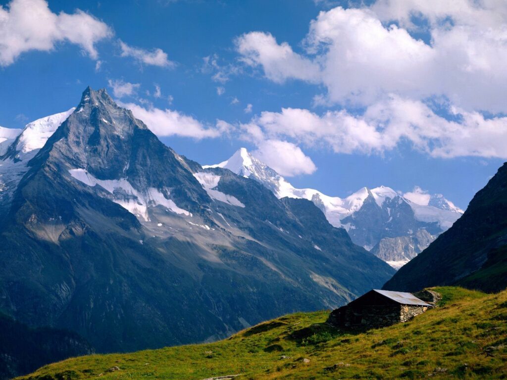 Snow-capped Swiss mountains with rustic house in green meadow under bright blue sky clouds Wallpaper