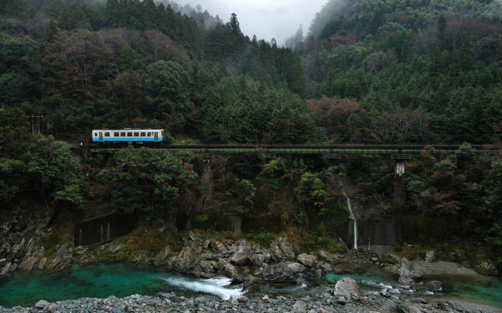 Train crossing narrow railroad bridge over turquoise river in misty forest hills wallpaper background