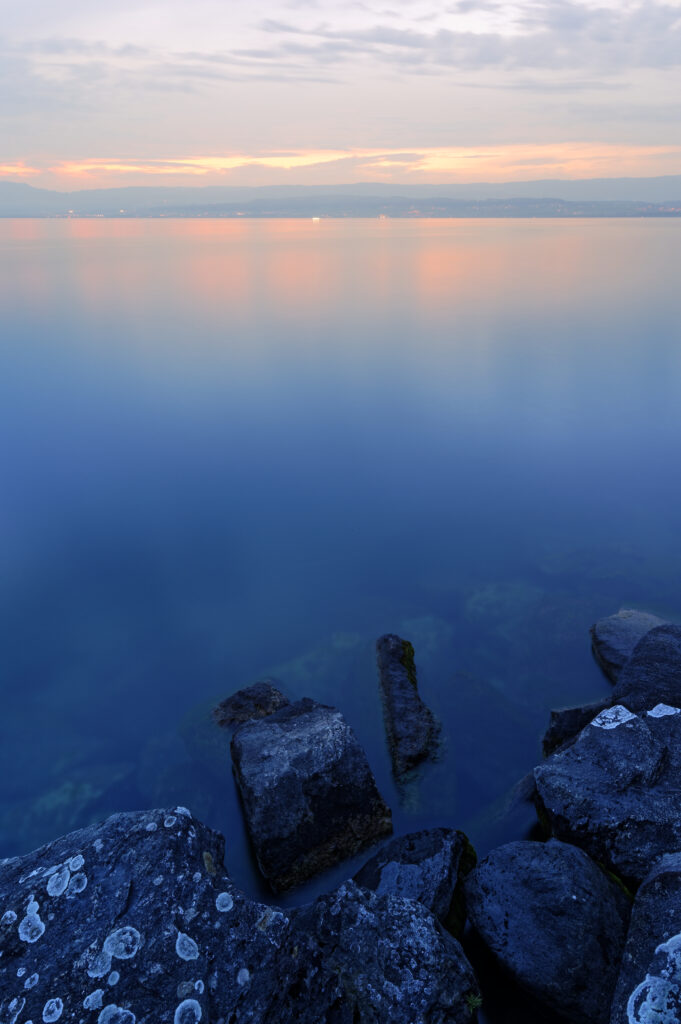 Tranquil lake with smooth water reflecting pastel sunset sky and dark stones in foreground Wallpaper