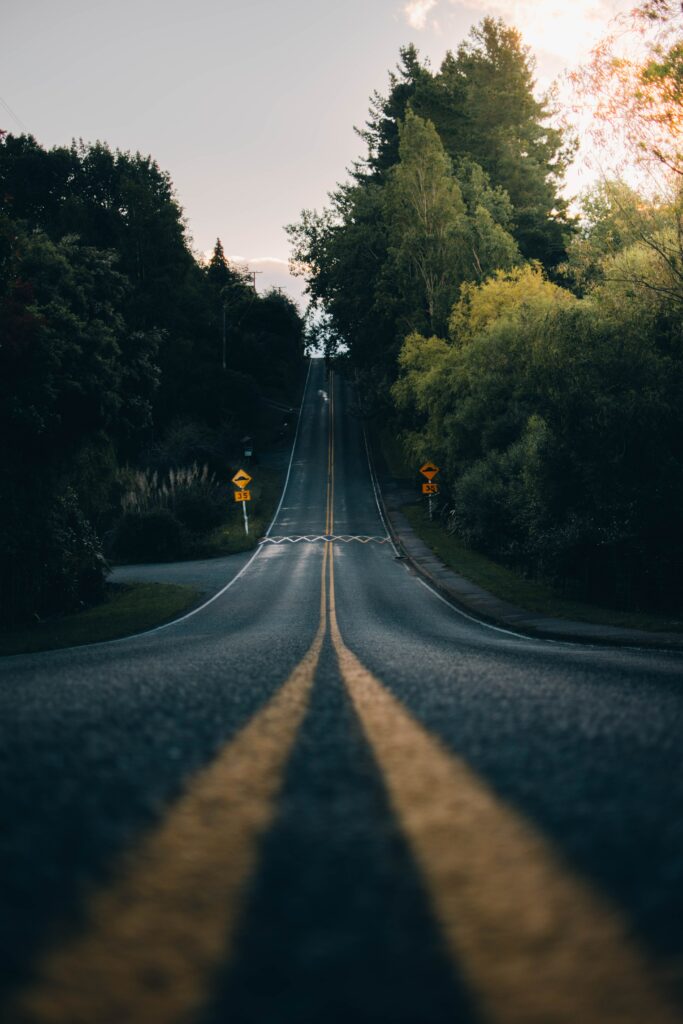 Serene road with lush green trees and vivid yellow center lines under soft evening light wallpaper