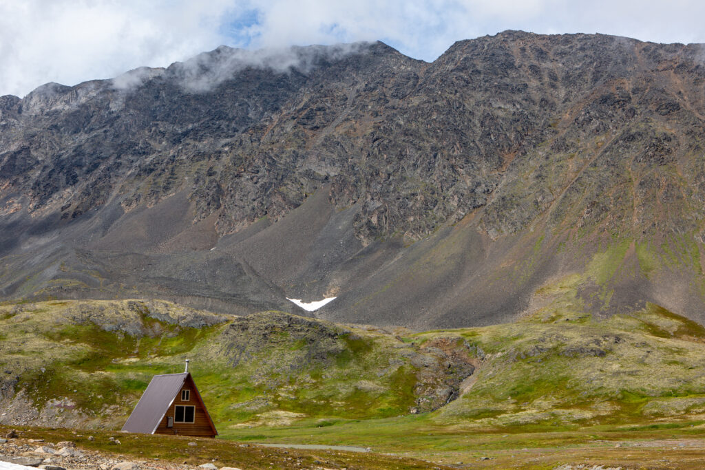Solitary triangular house in lush green grass below rocky hills with cloud-covered mountain peaks wallpaper