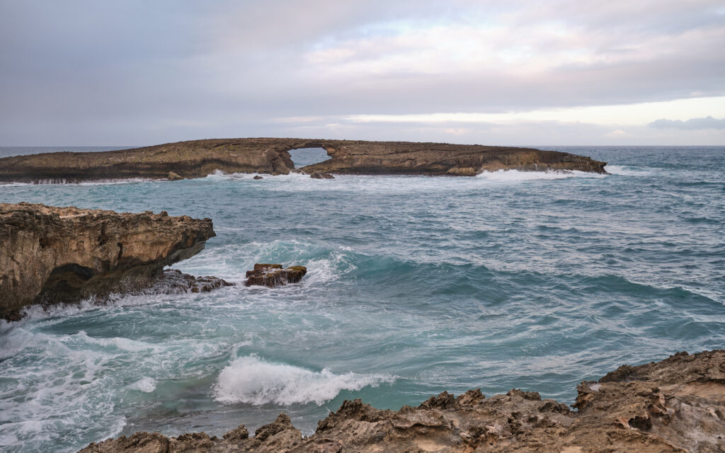 Serene turquoise sea waves hitting rocky cliffs with unique rock formation under cloudy sky Wallpaper