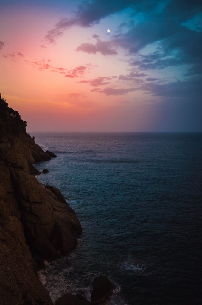 Serene twilight coastal wallpaper with calm sea rocks pastel sky pink orange blue hues moon rising